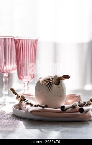 A stylish Easter dining setup with a focal decorative egg adorned with feathers, nestled on a pastel tablecloth, complemented by elegant pink-tinted g Stock Photo