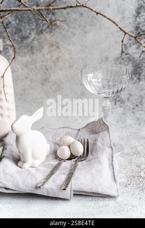 An elegant Easter-themed display featuring a white ceramic rabbit, three speckled eggs, and a clear glass. This setup is complemented by silverware an Stock Photo