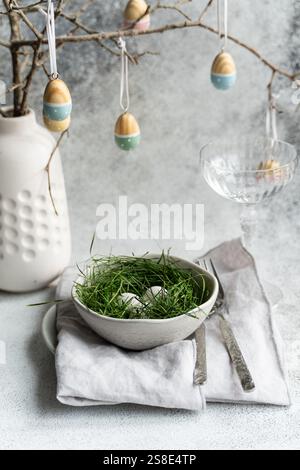 An elegantly set Easter table featuring a bowl of eggs nestled in fresh green grass, accompanied by a delicate arrangement of hanging artfully painted Stock Photo