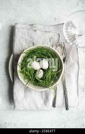 Top view of a simple yet elegant Easter table setting featuring a bowl with a nest of fresh green grass and three speckled eggs. A pair of vintage for Stock Photo