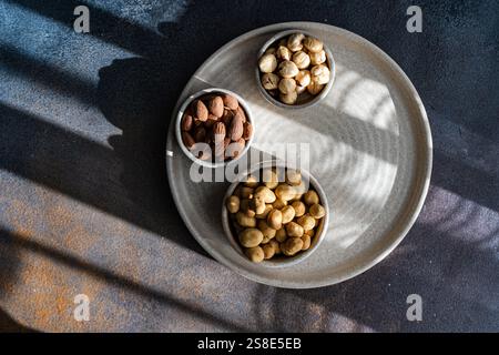 Top view of a circular plate holding three bowls filled with almonds, hazelnuts, and peanuts, beautifully highlighted by natural sunlight and shadows. Stock Photo