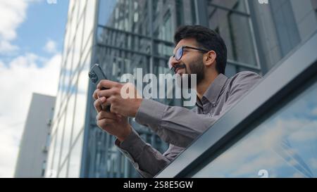 Smiling Arabian Indian man businessman entrepreneur manager use mobile phone application in city office terrace bottom view male business owner Stock Photo