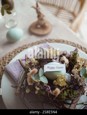 Top view of an elegantly arranged Easter table setting featuring naturally dyed eggs nestled in a decorative nest with spring flowers and a tag readin Stock Photo