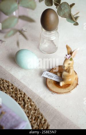 From above, an Easter table is set with naturally dyed eggs in soft pastel shades, accompanied by an ornamental bunny figure holding a Happy Easter si Stock Photo
