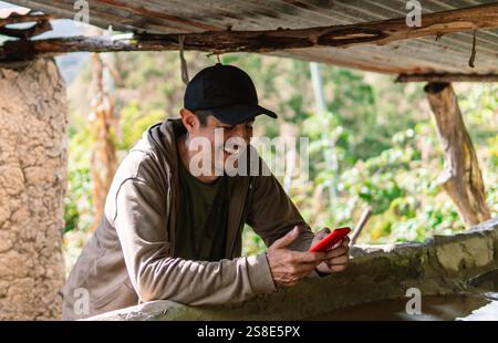 A man in a rural area of Colombia leans on a rustic structure, smiling while using a red smartphone Surrounded by greenery, he enjoys a moment of conn Stock Photo