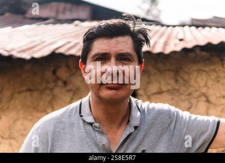 A close-up portrait of a man standing outdoors in a rural area of Colombia, with a rustic clay wall and corrugated metal roof in the background, refle Stock Photo