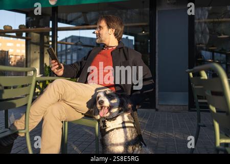 A man sits at a city cafe with his dog, browsing something on his smartphone in the warm sunlight Stock Photo