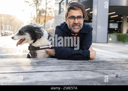 A middle-aged man enjoys a quiet moment with his black and white dog while sitting at a wooden table in an urban park. Both look leisurely and content Stock Photo