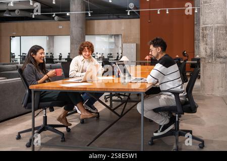 A diverse group of coworkers engaging in teamwork around a large wooden table in a modern office They discuss projects, share ideas, and utilize lapto Stock Photo
