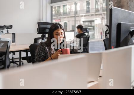 Diverse coworkers collaborating in a modern open-plan office A focused woman holds a mug while working, embodying teamwork, productivity, and a dynami Stock Photo