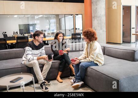 A group of diverse coworkers sits on a comfortable office sofa, engaging in a cheerful conversation over coffee The open-plan workspace fosters teamwo Stock Photo