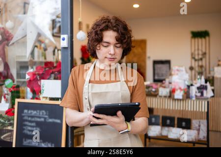 A young male employee stands in front of an organic produce store, wearing an apron and using a tablet to manage inventory The store is decorated with Stock Photo