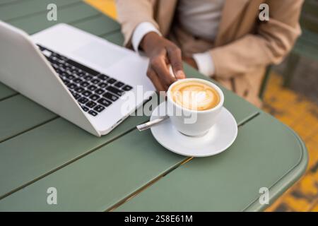Urban black woman working on a laptop while enjoying a cappuccino at an outdoor cafe A blend of technology and relaxation in a city setting exemplifie Stock Photo