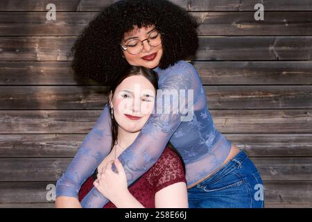 Two young Gen Z women, one Hispanic with curly hair, and the other of unspecified ethnicity, share a close hug. The Hispanic woman looks down while th Stock Photo