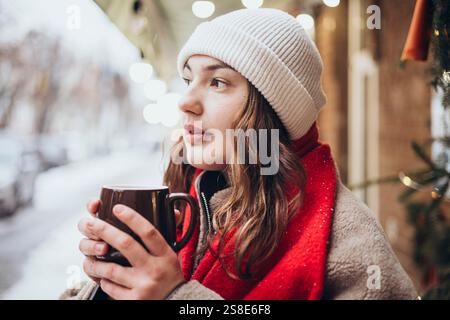 A young woman sips a hot chocolate on a snowy winter day. Dressed warmly in a white beanie and red scarf, she gazes thoughtfully while holding her mug Stock Photo