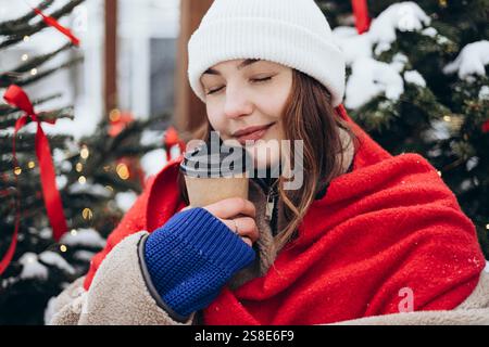 A young woman wrapped in a red scarf sips coffee near a Christmas tree on a snowy day, a moment of warmth and comfort. Stock Photo