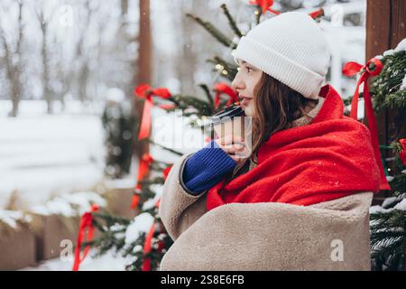 Young woman wrapped in a red scarf, holding a coffee cup, and smiling, seated near a festive Christmas tree on a snowy day. Stock Photo