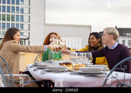 A joyful group of friends toasting with wine glasses on a rooftop The table is elegantly set, capturing a lively celebration with smiles and cheerful Stock Photo