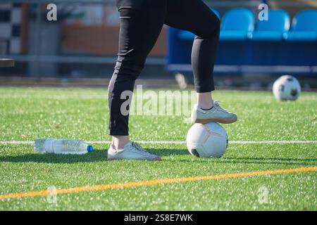A person wearing sneakers and black pants stands with one foot on a soccer ball on a grass soccer field. The scene includes another ball and a water b Stock Photo