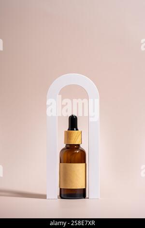 An amber glass dropper bottle with a wooden cap and blank label is elegantly placed in a white arch frame against a beige background, highlighting min Stock Photo