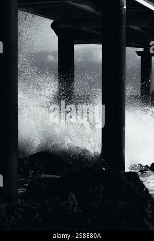 Black and white photo captures powerful waves crashing against a pier's pillars. The scene highlights the raw natural beauty and dynamic energy of Lan Stock Photo