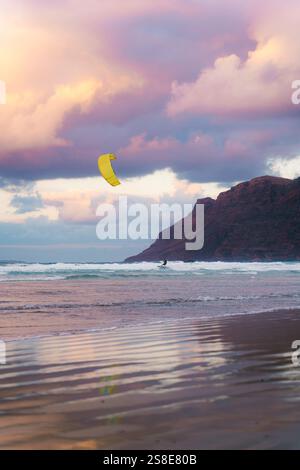 A kite surfer glides across the waves at Famara Beach, Lanzarote, under a sky painted with stunning sunset hues. The dramatic cliffs add to the breath Stock Photo