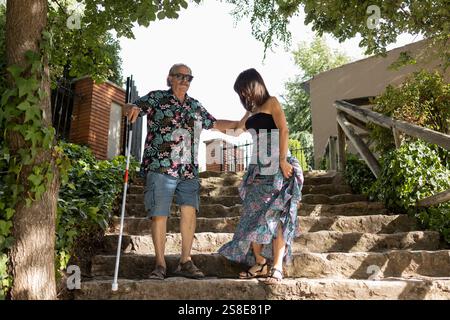 A blind elderly man with sunglasses and a white cane is guided gently by his adult daughter down a flight of stone steps amidst green surroundings. Stock Photo