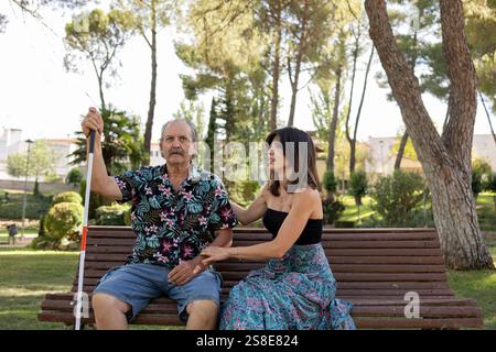 A blind father with a disability sitting next to his daughter on a park bench, holding a white cane, as she lovingly assists him. Stock Photo