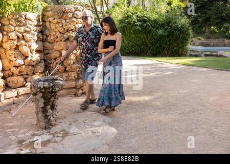 A blind man with a white cane is carefully guided by his supportive daughter through a picturesque park, showcasing a touching moment of family care a Stock Photo