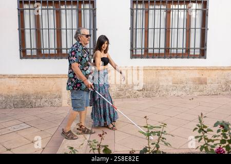 A daughter assists her blind father, who uses a white cane, as they walk together past a building with large windows and a stone facade. Stock Photo