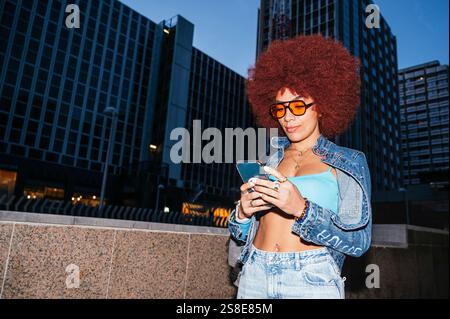 African descendent woman with afro hair, wearing orange glasses, uses her smartphone against an urban backdrop. She is stylishly dressed in a denim ja Stock Photo