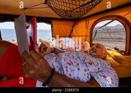 Gay senior couple enjoying a leisurely day inside their off-road camper, looking at a tablet with the ocean in the background. They are relaxed and co Stock Photo