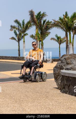 A man in a wheelchair is captured enjoying the sunny weather on a paved path. He is dressed in a casual yellow shirt and black shorts, with sunglasses Stock Photo