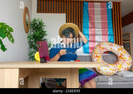 Transgender man hosting a virtual meeting from home, displayed on a smartphone. He is sitting at a wooden table, wearing a straw hat and a blue shirt. Stock Photo