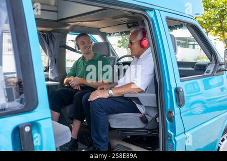 A young man and an elderly man in a wheelchair share a joyful moment inside a blue camper van. The younger man, seated in the driver's seat, is attent Stock Photo