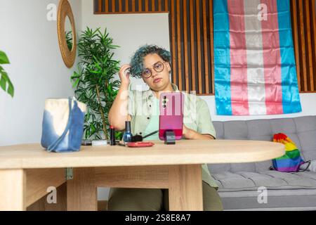 A transgender man sits at a wooden table, engaging in a makeup tutorial using his mobile phone. With pride flag in the background, he is surrounded by Stock Photo
