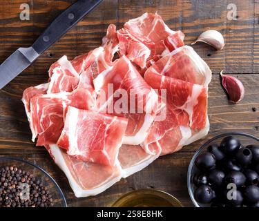 Thinly sliced prosciutto arranged on a wooden board, accompanied by black olives and a bowl of olive oil. A rustic setting enhances the gourmet appeal Stock Photo