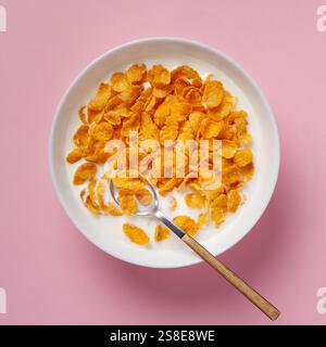 Top view of a white bowl filled with golden cornflakes and milk, accompanied by a metal spoon, set against a soft pink background. Perfect for a healt Stock Photo