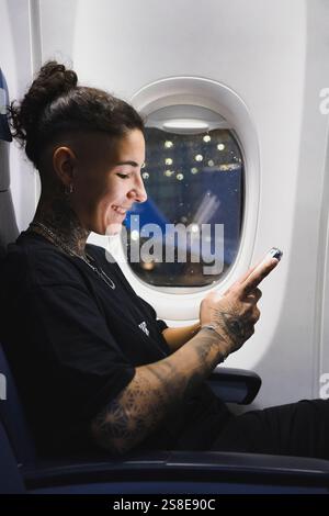 A young woman with intricate tattoos sits on an airplane, smiling while using a smartphone The window reveals a beautiful night, and the ambient light Stock Photo