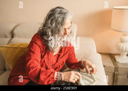 Elderly woman with gray hair sits on a beige couch, focused on sewing. A warm lamp light casts a soft ambiance in a cozy living room setting, enhancin Stock Photo