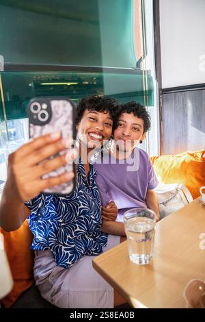 A joyful, multiethnic LGBTQ+ couple sits in a cozy cafe, smiling as they take a selfie. Sunlight streams through the window, illuminating their happy Stock Photo