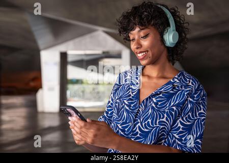 A woman wearing blue headphones smiles while looking at her phone, standing in a modern architectural space. She wears a blue and white patterned shir Stock Photo