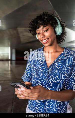 Woman with curly hair wearing blue patterned shirt listens to music on headphones while smiling and looking at her smartphone in a modern indoor setti Stock Photo