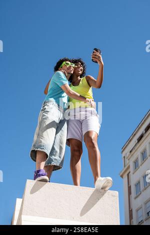 A joyful, multiethnic LGBTQ+ couple captures a selfie outdoors. They stand closely on a sunny day, showcasing unity and happiness against a clear blue Stock Photo
