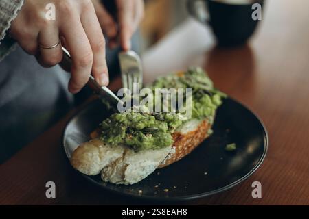 A person slices avocado toast topped with seeds on a dark plate. The focus is on the dining experience, with a blurred background, highlighting a warm Stock Photo