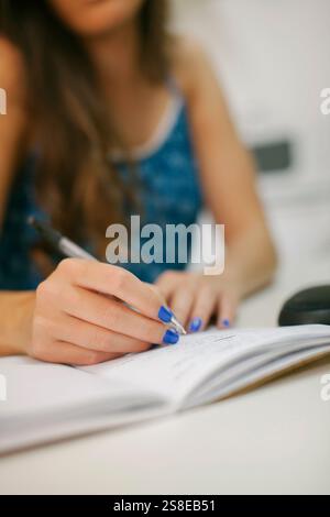 A woman with bright blue nails is writing in a notebook. The focus is on her hand holding the pen, emphasizing concentration and creativity in a works Stock Photo