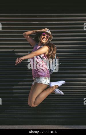 A young woman in sunglasses and a hat jumps joyfully against a dark urban background, embodying freedom and happiness. Perfect for lifestyle and youth Stock Photo