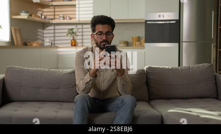 Arabian Indian ethnic man guy use mobile phone on couch browsing mobile internet at home kitchen take break confused read bad news on smartphone male Stock Photo