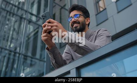 Smiling Arabian Indian man businessman entrepreneur manager use mobile phone application in city office terrace bottom view male business owner Stock Photo