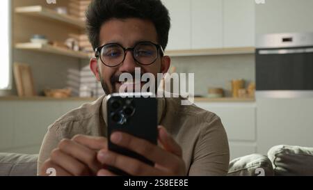 Arabian Indian man guy happy smiling male freelancer relaxing on couch sofa at home kitchen use mobile phone gadget scrolling social media smartphone Stock Photo